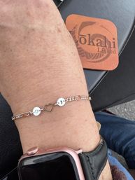 Close-up of a wrist wearing a delicate silver chain bracelet with two round initial discs 'D' and 'J' flanking a heart-shaped charm, paired with a pink metal-cased smartwatch against a black leather surface.