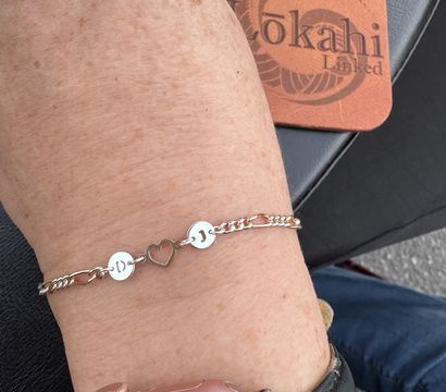 Close-up of a wrist wearing a delicate silver chain bracelet with two round initial discs 'D' and 'J' flanking a heart-shaped charm, paired with a pink metal-cased smartwatch against a black leather surface.