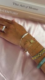 Close-up of a hand on pink satin wearing stacked gold chain bracelets with a coin charm, a beaded bangle, two-tone metal bracelet, jade bangle and a diamond ring, books in background.