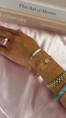 Close-up of a hand on pink satin wearing stacked gold chain bracelets with a coin charm, a beaded bangle, two-tone metal bracelet, jade bangle and a diamond ring, books in background.