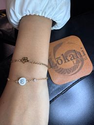 Close-up of a wrist on a manicure table wearing two delicate gold chain bracelets — a flower charm and a mother-of-pearl monogram disc with letter E and a small crystal, beside a brown leather coaster with a circular logo.