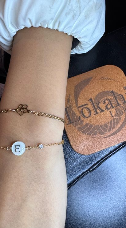 Close-up of a wrist on a manicure table wearing two delicate gold chain bracelets — a flower charm and a mother-of-pearl monogram disc with letter E and a small crystal, beside a brown leather coaster with a circular logo.