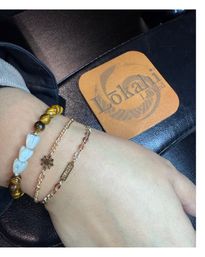 Close-up of a wrist with stacked bracelets — tiger's eye bead bracelet with white shell-like beads and two delicate gold chain bracelets with a tiny sun charm and small engraved tag — resting on a black leather wallet with a tan logo patch.