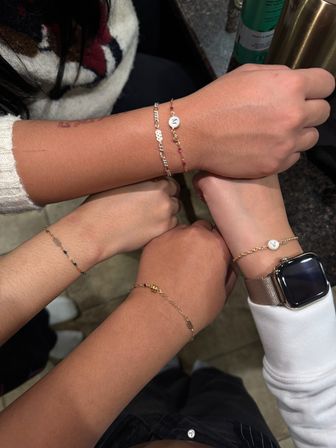 Close-up of four wrists joined over a countertop wearing delicate gold and beaded bracelets, two with round initial coin charms and one wrist with a metal mesh smartwatch, friendship-style jewelry shot.