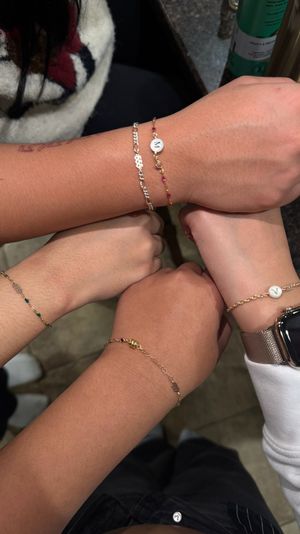 Close-up of four wrists joined over a countertop wearing delicate gold and beaded bracelets, two with round initial coin charms and one wrist with a metal mesh smartwatch, friendship-style jewelry shot.