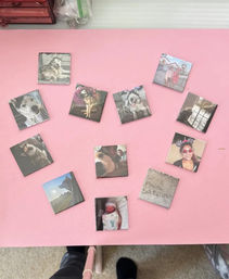 Overhead view of small square photo magnets arranged in a heart on a pink surface — pictures of husky dogs, a newborn, selfies and scenic shots; photographer’s legs and selfie stick visible at the bottom.