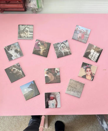 Overhead view of small square photo magnets arranged in a heart on a pink surface — pictures of husky dogs, a newborn, selfies and scenic shots; photographer’s legs and selfie stick visible at the bottom.