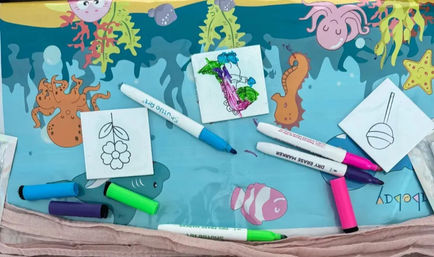 Bright ocean-themed kids' craft table with small tile coloring sets and scattered neon dry-erase markers on a blue play mat decorated with octopus, seahorse and clownfish; pricing cards and photo prints peek at the bottom.