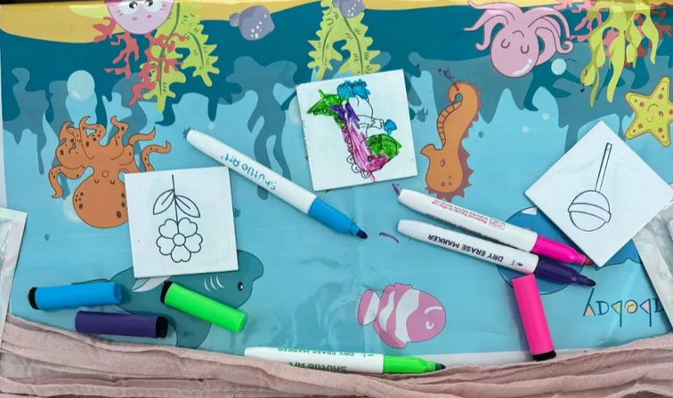 Bright ocean-themed kids' craft table with small tile coloring sets and scattered neon dry-erase markers on a blue play mat decorated with octopus, seahorse and clownfish; pricing cards and photo prints peek at the bottom.