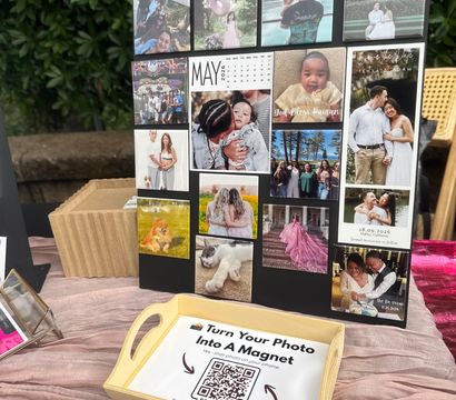 Colorful photo magnets on a black display board at an outdoor market table — family, baby, pet and wedding images with a wooden tray holding a QR sign reading ‘Turn Your Photo Into A Magnet’, pink tablecloth and leafy backdrop.