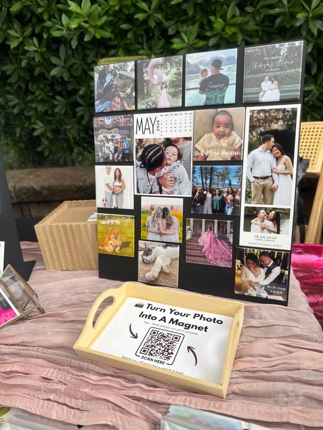 Colorful photo magnets on a black display board at an outdoor market table — family, baby, pet and wedding images with a wooden tray holding a QR sign reading ‘Turn Your Photo Into A Magnet’, pink tablecloth and leafy backdrop.
