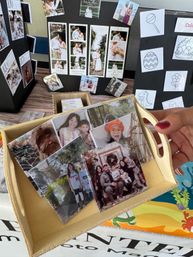 Wooden tray of glossy custom photo magnets and tiles featuring family portraits and kids, held by a hand at a craft‑market table with black display boards showing photo strips and save‑the‑date samples.