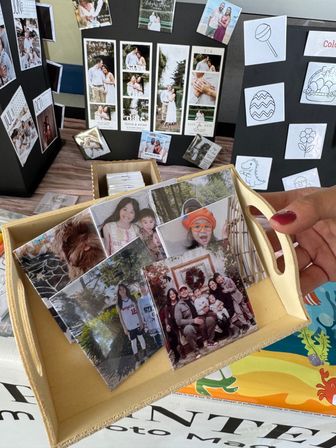 Wooden tray of glossy custom photo magnets and tiles featuring family portraits and kids, held by a hand at a craft‑market table with black display boards showing photo strips and save‑the‑date samples.