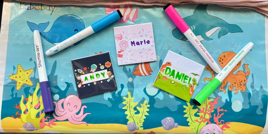 Overhead view of a kids' craft table with an underwater-themed placemat featuring fish, octopus and starfish, three small personalized square name tiles, and five colorful dry-erase markers scattered across it.