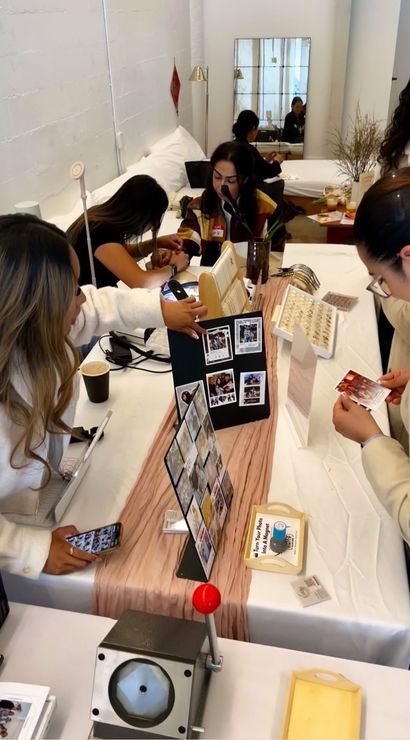 Pop-up photo booth in a bright studio: attendees gathered around a table of instant photos and DIY magnet displays, with photo grids, instant prints, a small red-lever magnet press, jewelry trays, phones and coffee