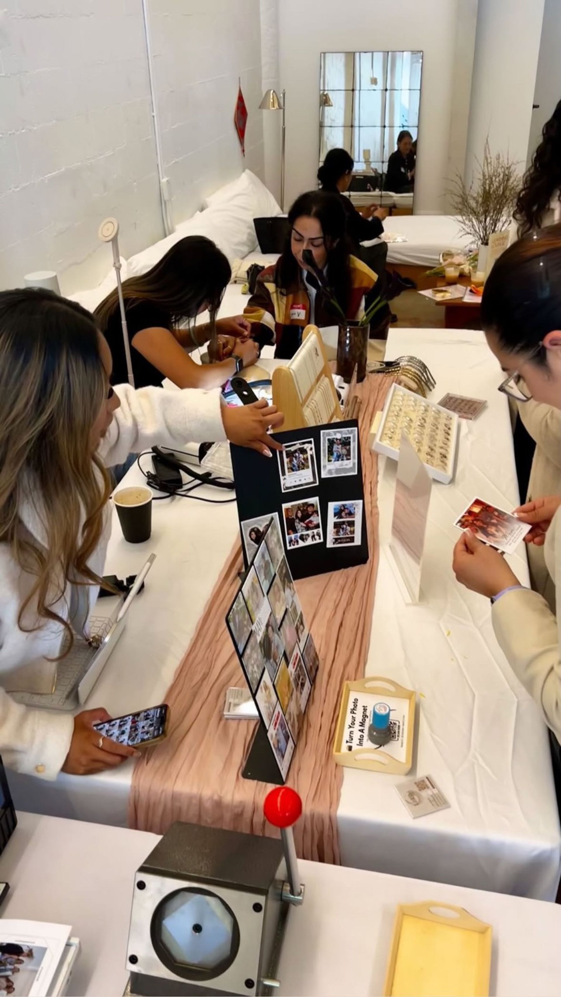 Pop-up photo booth in a bright studio: attendees gathered around a table of instant photos and DIY magnet displays, with photo grids, instant prints, a small red-lever magnet press, jewelry trays, phones and coffee