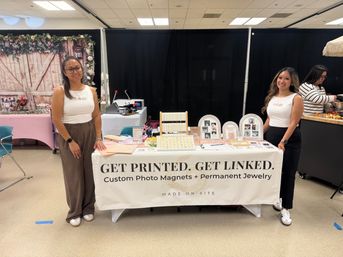 Two women at an indoor vendor booth offering custom photo magnets and permanent jewelry, with framed photo samples, jewelry display racks and a floral backdrop