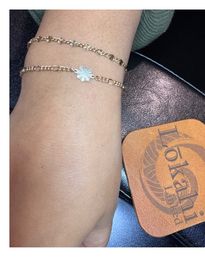 Close-up of a wrist wearing two dainty gold chain bracelets—one with tiny floral links and one with a small sunburst charm—resting on a dark leather surface beside a tan leather coaster.