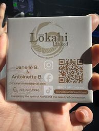 Close-up of a hand holding a small jewelry product box with a circular logo, QR code, social media icons and the tagline “Inspired by the spirit of Aloha,” suggesting Hawaii‑inspired jewelry packaging.