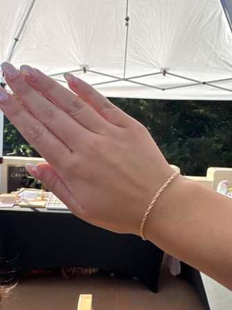 Close-up of a hand with glossy pink ombré nails wearing a delicate twisted gold chain bracelet beneath a white canopy at an outdoor market stall.