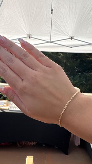 Close-up of a hand with glossy pink ombré nails wearing a delicate twisted gold chain bracelet beneath a white canopy at an outdoor market stall.