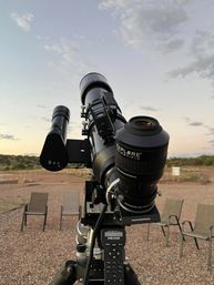 Close-up of a stargazing telescope on a tripod at a desert observing site at dusk, eyepiece and finder scope in focus with gravel ground and empty folding chairs beneath a pastel sky.