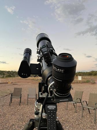 Close-up of a stargazing telescope on a tripod at a desert observing site at dusk, eyepiece and finder scope in focus with gravel ground and empty folding chairs beneath a pastel sky.