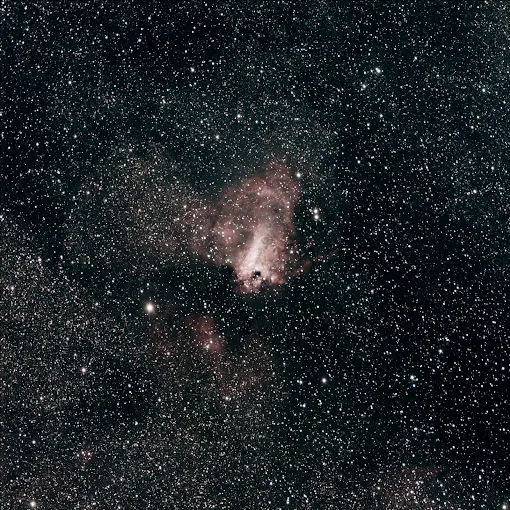 Cosmic deep-sky view: dense Milky Way star field with a pinkish emission nebula and dark dust lanes against black space.