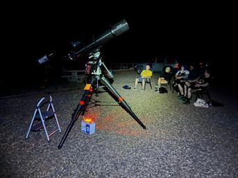 Large equatorial telescope on tripod with red safety light on a gravel lot at night, a small group in folding chairs nearby enjoying stargazing