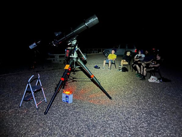 Large equatorial telescope on tripod with red safety light on a gravel lot at night, a small group in folding chairs nearby enjoying stargazing