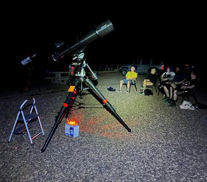 Large equatorial telescope on tripod with red safety light on a gravel lot at night, a small group in folding chairs nearby enjoying stargazing