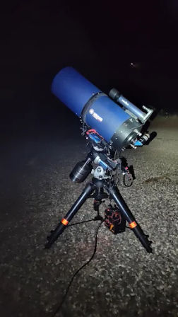 Large blue telescope on an equatorial mount and tripod set up on gravel at night, cables and counterweights visible, ready for stargazing/astrophotography under a dark sky