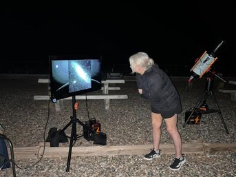 Curious stargazer leaning in to view a galaxy image on a monitor during an outdoor night-sky session, with a telescope on a tripod, portable power packs, and picnic tables on a gravel area under a dark sky.