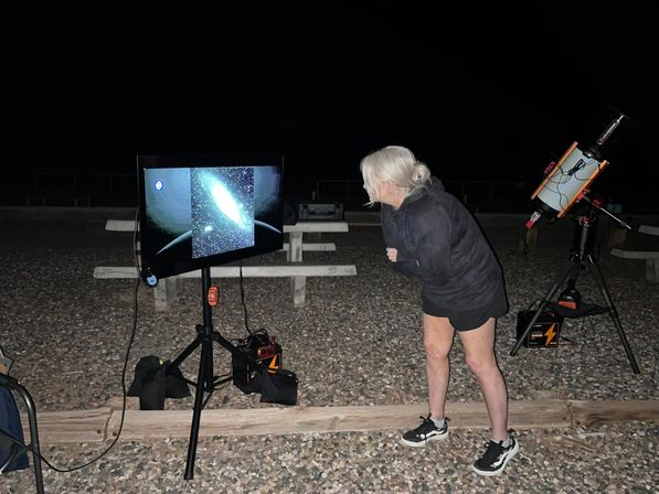 Curious stargazer leaning in to view a galaxy image on a monitor during an outdoor night-sky session, with a telescope on a tripod, portable power packs, and picnic tables on a gravel area under a dark sky.