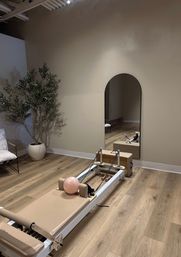 Pilates reformer in a minimalist Pilates studio with a pale pink exercise ball, arched wall mirror, potted plant, and light wood floors.
