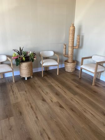 Minimalist reception waiting area with three light wood-framed beige chairs, a large wicker planter of mixed tropical flowers, a whimsical woven cactus sculpture in a basket, and warm wood-look plank flooring against neutral beige walls.