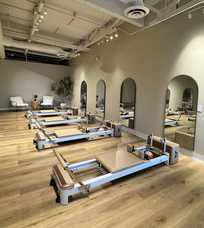 Minimalist Pilates studio interior with multiple reformer machines in a row on light wood floors, arched wall mirrors, neutral decor, potted plant and lounge seating — bright modern fitness space.