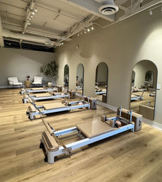 Minimalist Pilates studio interior with multiple reformer machines in a row on light wood floors, arched wall mirrors, neutral decor, potted plant and lounge seating — bright modern fitness space.