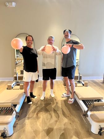 Three men playfully holding pink exercise balls in a bright pilates reformer studio with wooden floors, arched mirrors and reformer machines.