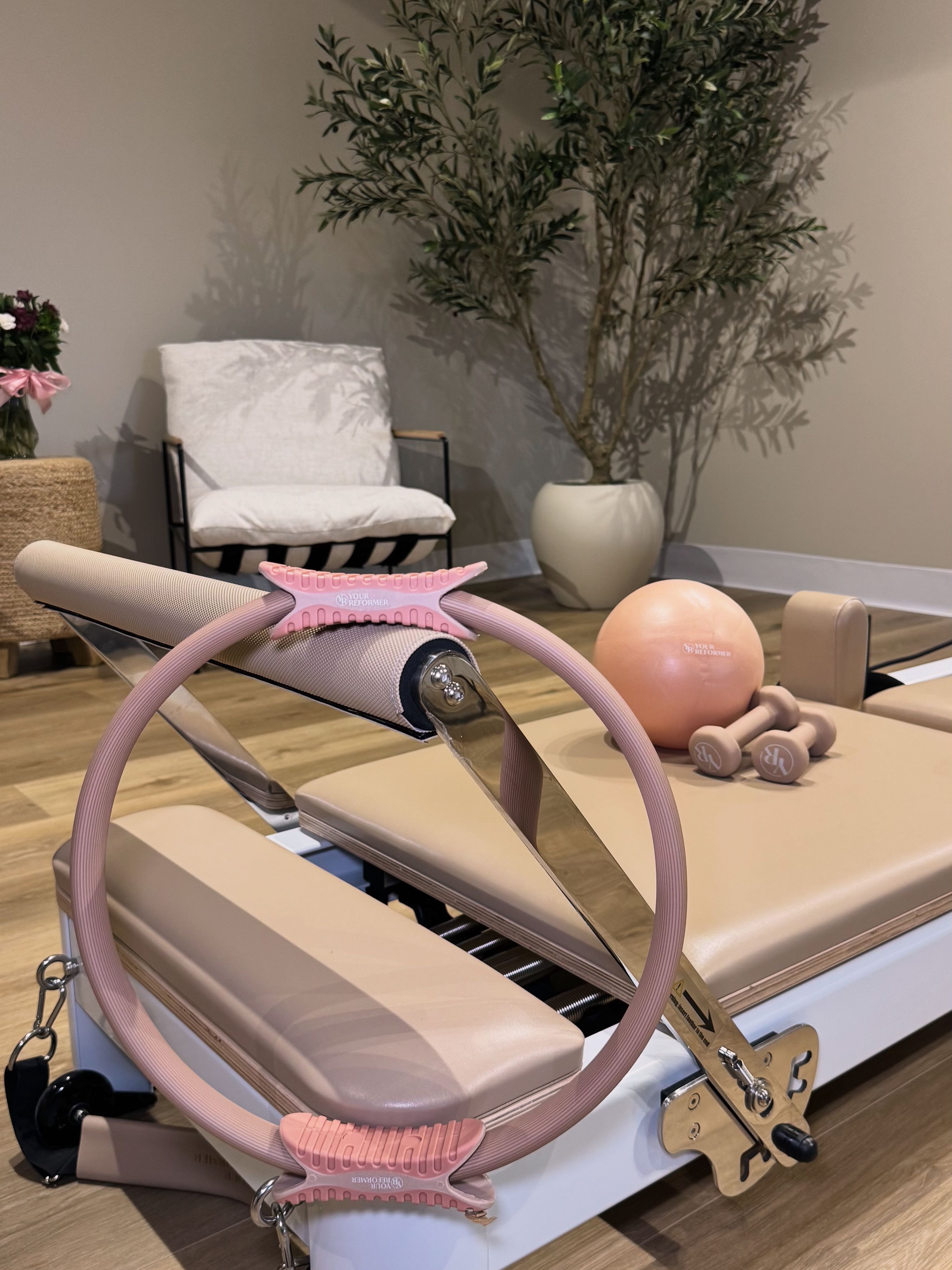 Pilates reformer with pink Pilates ring, mini exercise ball and light dumbbells on a beige carriage in a cozy neutral-toned Pilates studio with a chair and potted plant.