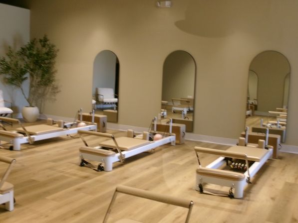 Bright, airy modern Pilates studio with multiple reformer machines on light wood floors, arched wall mirrors, neutral beige walls and a potted plant