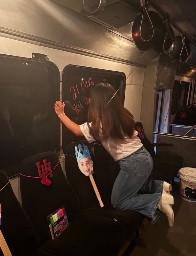 Dimly lit party bus interior with a person kneeling on a seat writing on a tinted window in orange marker; decorated seats, a photo cutout on a stick wearing a blue paper crown, and colorful markers tucked into a seat pocket.