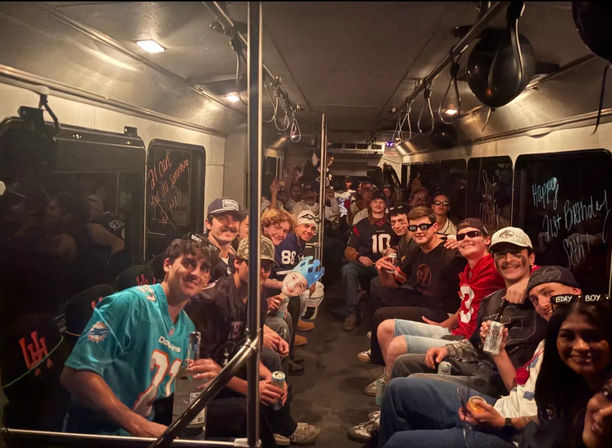 Large group of young adults partying inside a dimly lit party bus, seated along both sides in football jerseys and casual clothes, holding drinks, with birthday messages on the windows and one person wearing a blue crown.