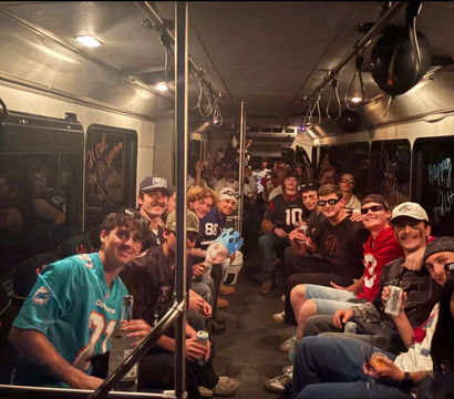 Large group of young adults partying inside a dimly lit party bus, seated along both sides in football jerseys and casual clothes, holding drinks, with birthday messages on the windows and one person wearing a blue crown.