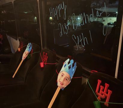 Nighttime party shuttle interior with black seats decorated for a birthday: smiling paper face cutouts on sticks wearing blue paper crowns, red-beaded garlands and small colored cups, and a festive 'Happy 21st Birthday' message written on the window.