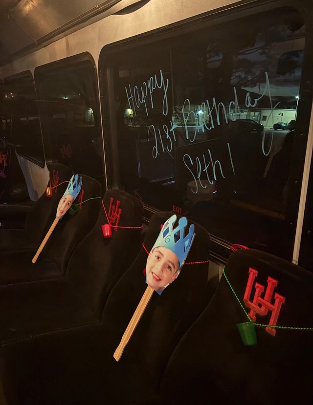 Nighttime party shuttle interior with black seats decorated for a birthday: smiling paper face cutouts on sticks wearing blue paper crowns, red-beaded garlands and small colored cups, and a festive 'Happy 21st Birthday' message written on the window.
