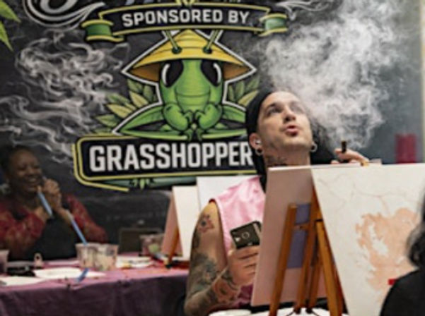 Person at an indoor painting event holding a phone and exhaling a large cloud of smoke beside an easel and canvas, seated in front of a colorful green grasshopper mural.