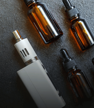 Flatlay of a sleek white box vape mod with amber glass e‑liquid dropper bottles on a dark textured surface, vaping accessories and e‑liquid bottles.