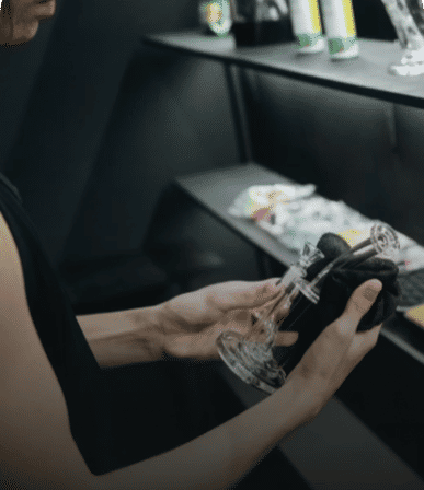 Hands polishing a stemmed cocktail glass with a black cloth behind a sleek dark bar shelf stocked with canned drinks and napkins