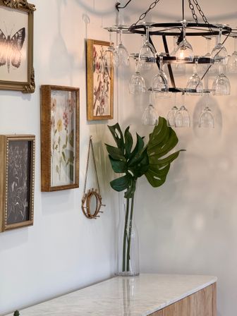 Stylish home interior: marble-topped console with tall glass vase of monstera leaves, vintage framed botanical prints on wall, and playful wine-glass chandelier.